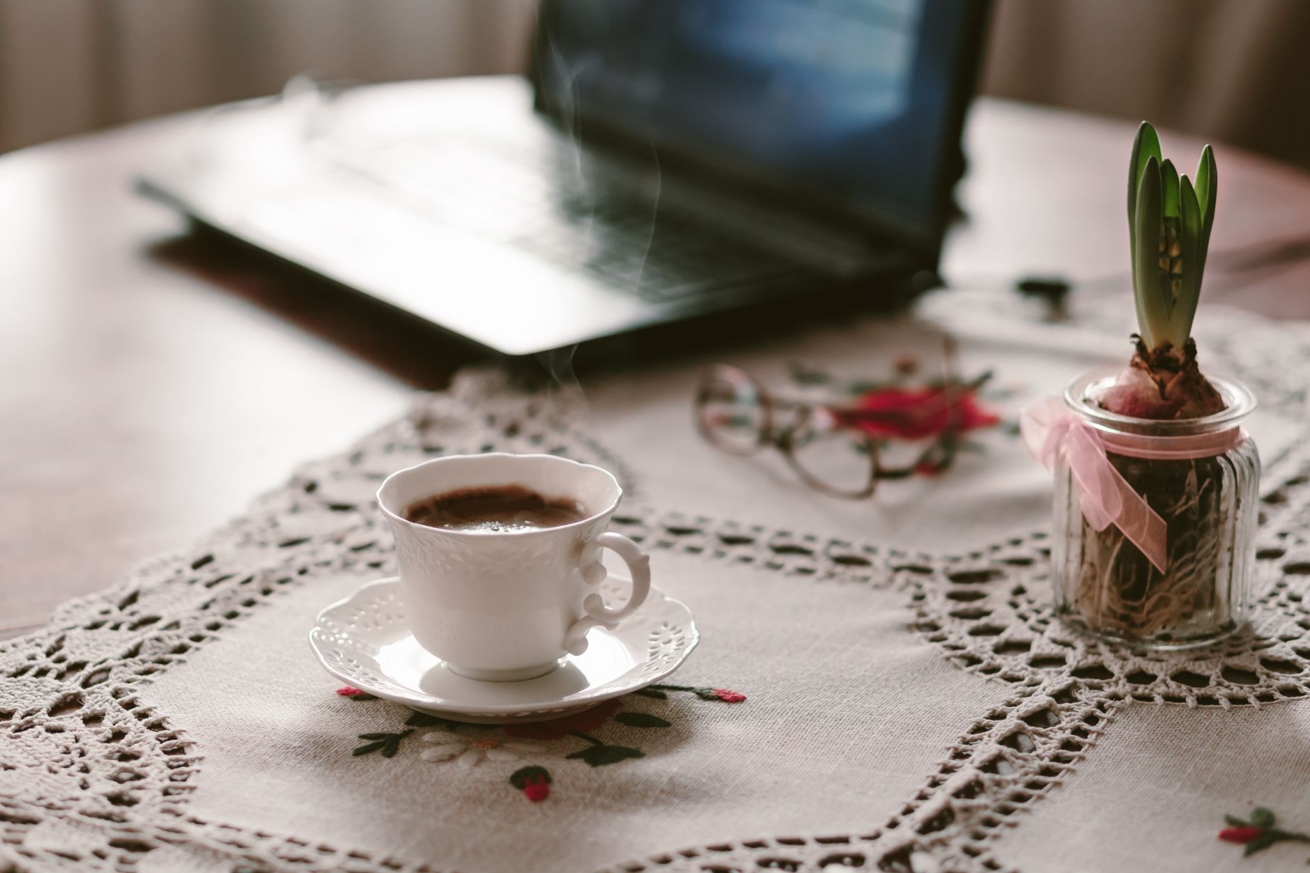 Ein einladender Arbeitsplatz zuhause mit einer dampfenden Tasse Kaffee auf einer Spitzendecke neben einem Laptop.